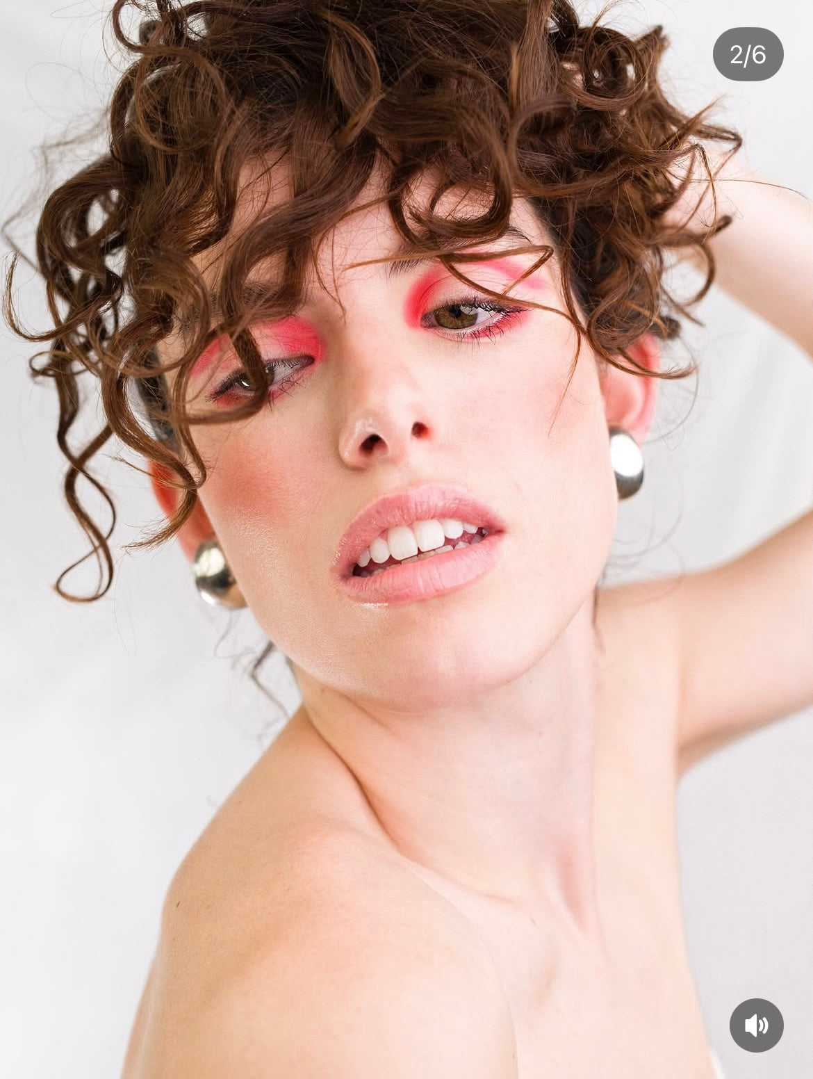 A close-up portrait of a woman with short curly brown hair, wearing dramatic pink and red eyeshadow, blush, and nude lipstick, with white pearl-like earrings against a light neutral background.